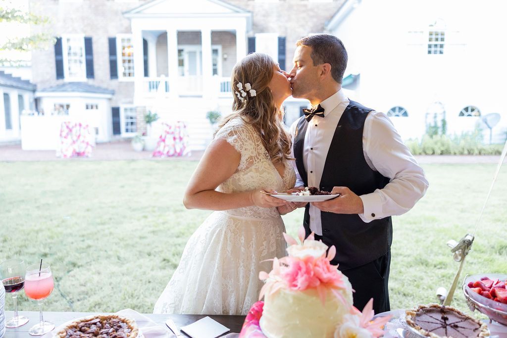 A couple in wedding attire kisses while holding dessert plates in front of a white building on a grassy lawn.