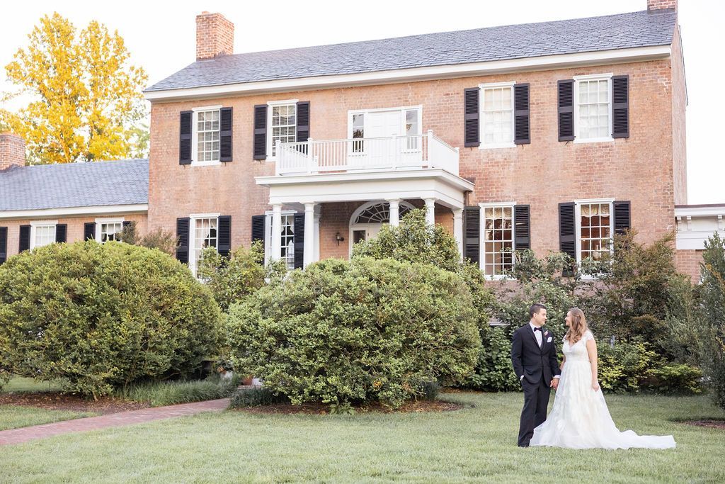 A couple in formal wedding attire stands on a lawn in front of a historic, two-story brick mansion with black shutters.