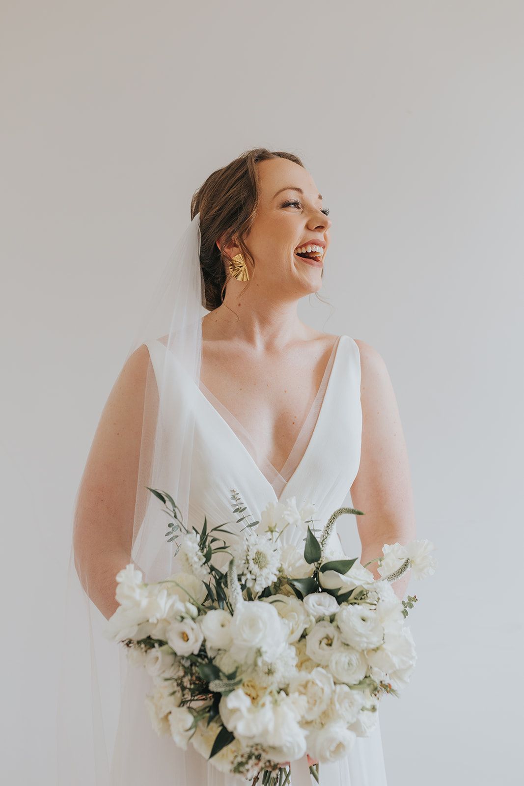 A bride smiling and holding a large bouquet of white flowers while wearing a wedding gown and veil against a plain wall.