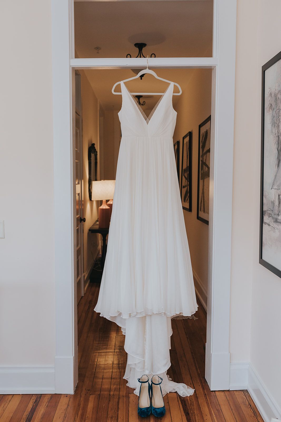 A white wedding dress hangs in a doorway above a pair of blue heels on a wooden floor in a home hallway.