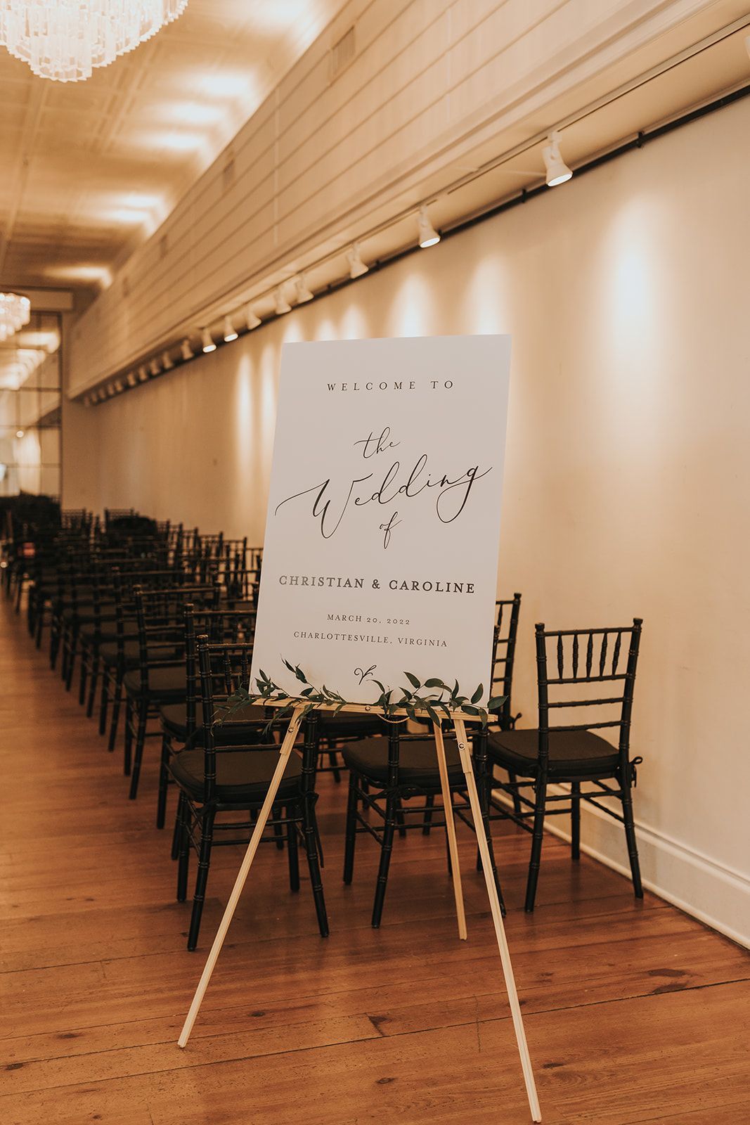 A welcome sign on an easel stands at the entrance of a venue hall with rows of chairs set up for a wedding ceremony.