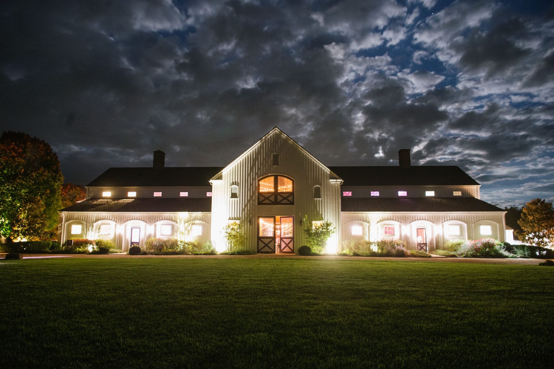A large, white, illuminated building with a central gable and barn-style features stands at night under a cloudy sky.