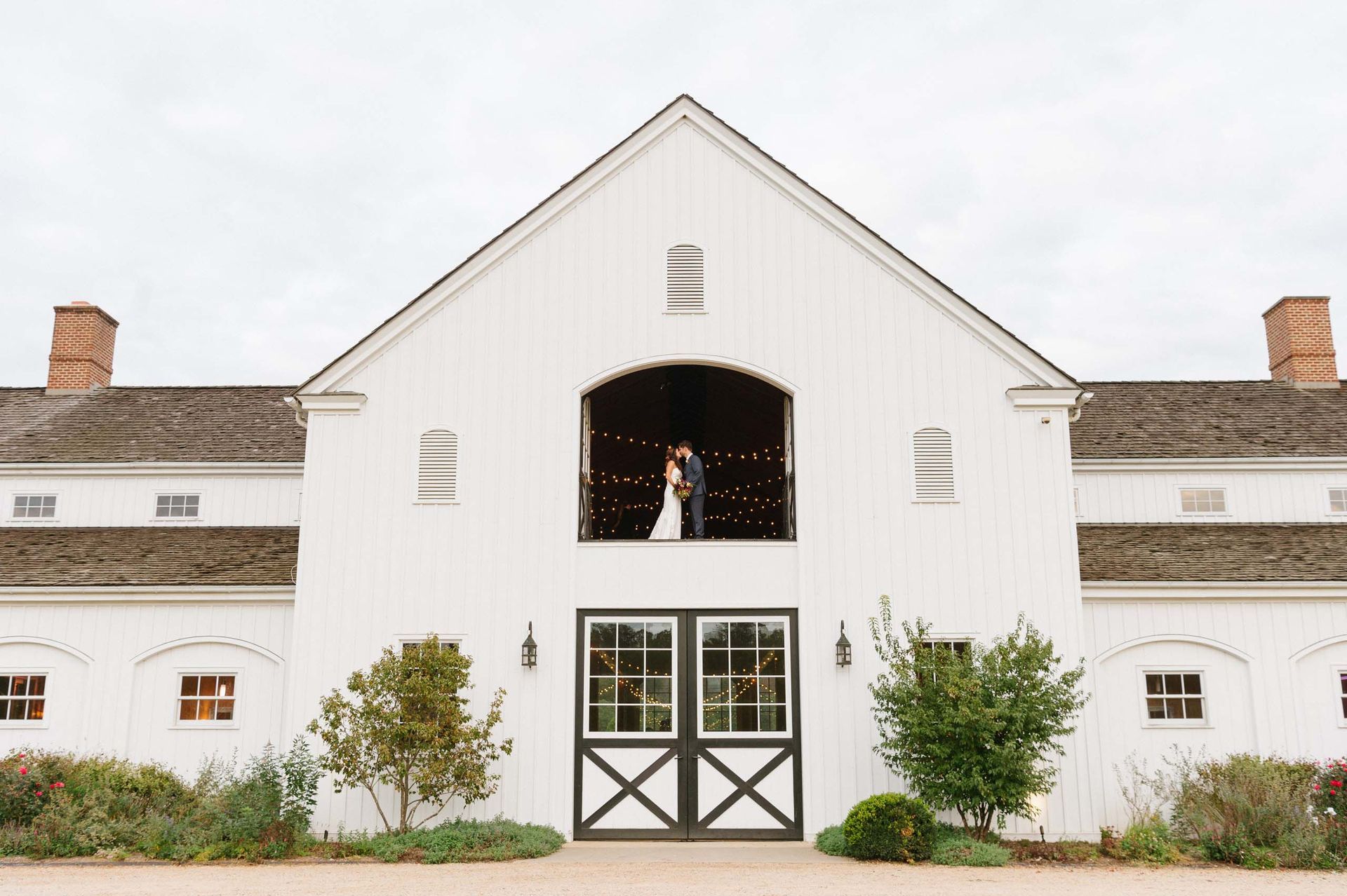 A couple stands inside the open upper balcony of a white wooden barn with a dark roof and large double doors.