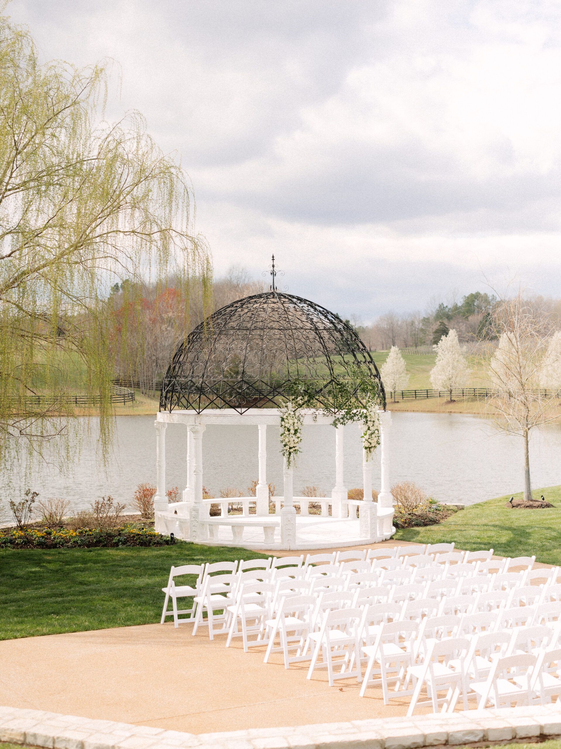 A white gazebo stands beside a pond, with rows of white chairs set up for an outdoor wedding ceremony.