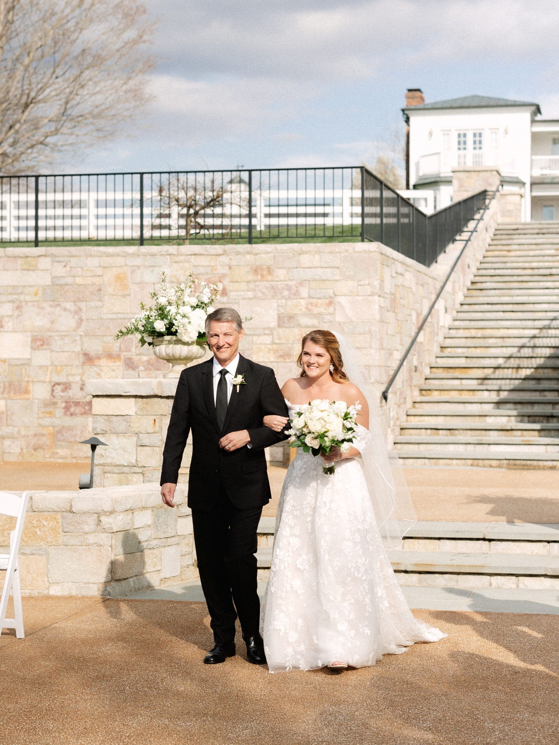A bride and a man in a tuxedo walk arm-in-arm outdoors in front of a stone wall, large staircase, and white building.