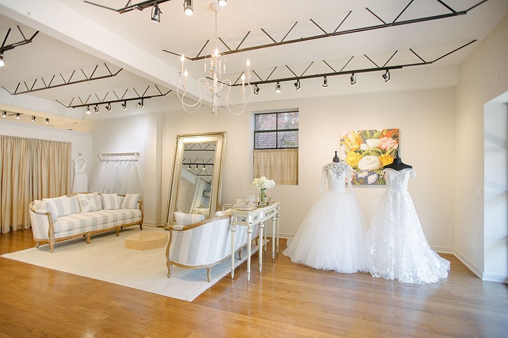A bright bridal boutique with a seating area, a large floor mirror, and two wedding dresses on mannequins.