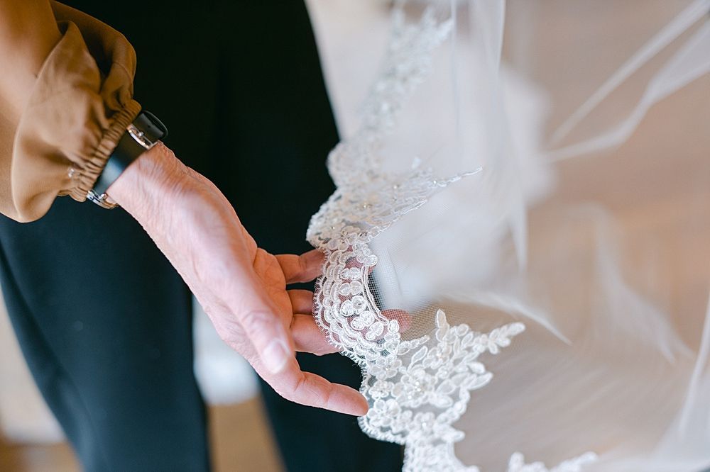 A hand holds up a delicate, lace-trimmed wedding veil, showcasing its intricate floral embroidery and beadwork details.