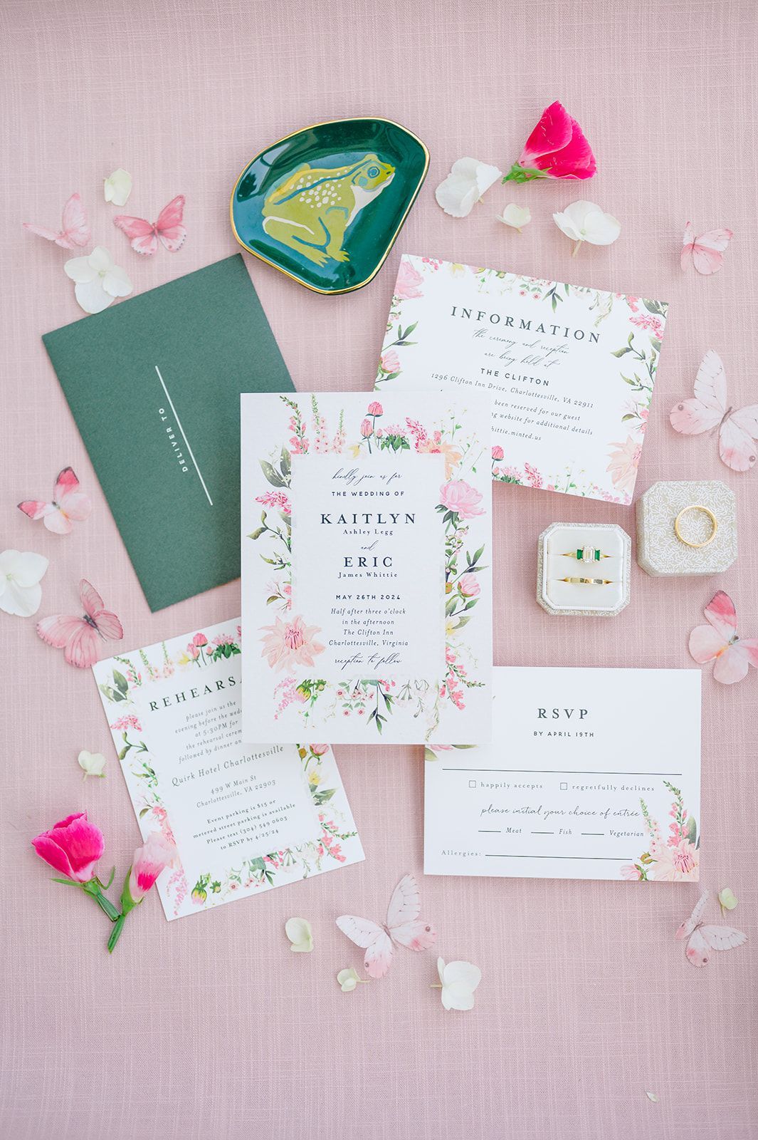 Flat lay of floral wedding invitations and envelopes in shades of pink and green, accented with butterflies and rings.