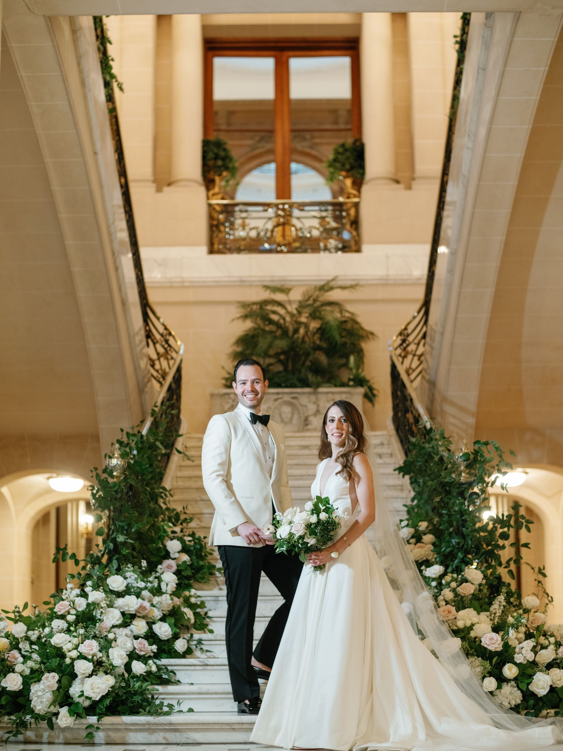 A couple in formal wedding attire stands on grand stairs adorned with white flowers and greenery inside a classic hall.