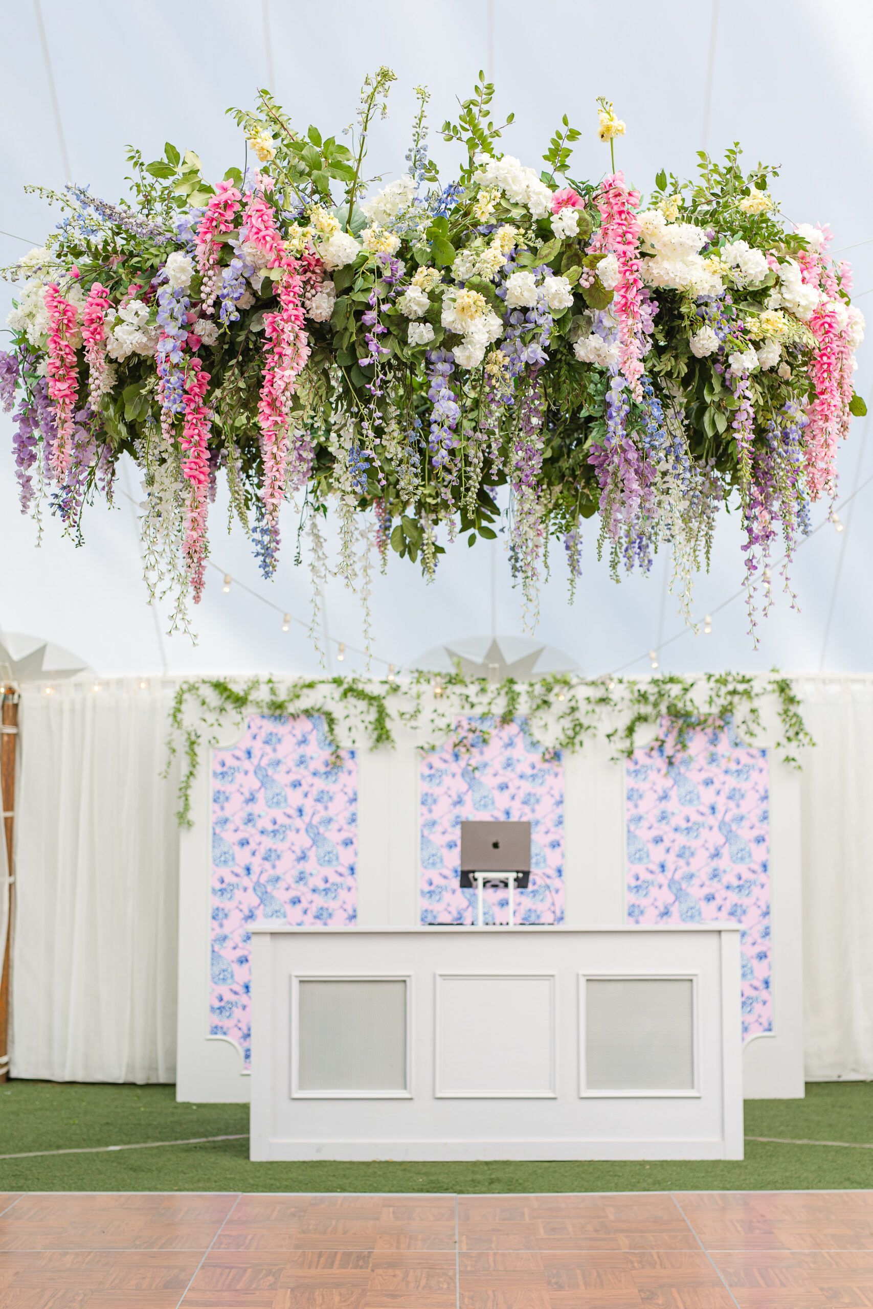 A white DJ booth sits before a floral-patterned wall, beneath a large, hanging chandelier of pink, blue, and white flowers.