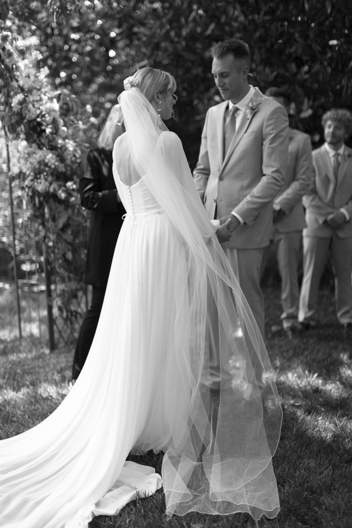 A couple stands outdoors during a wedding ceremony, holding hands while facing each other.