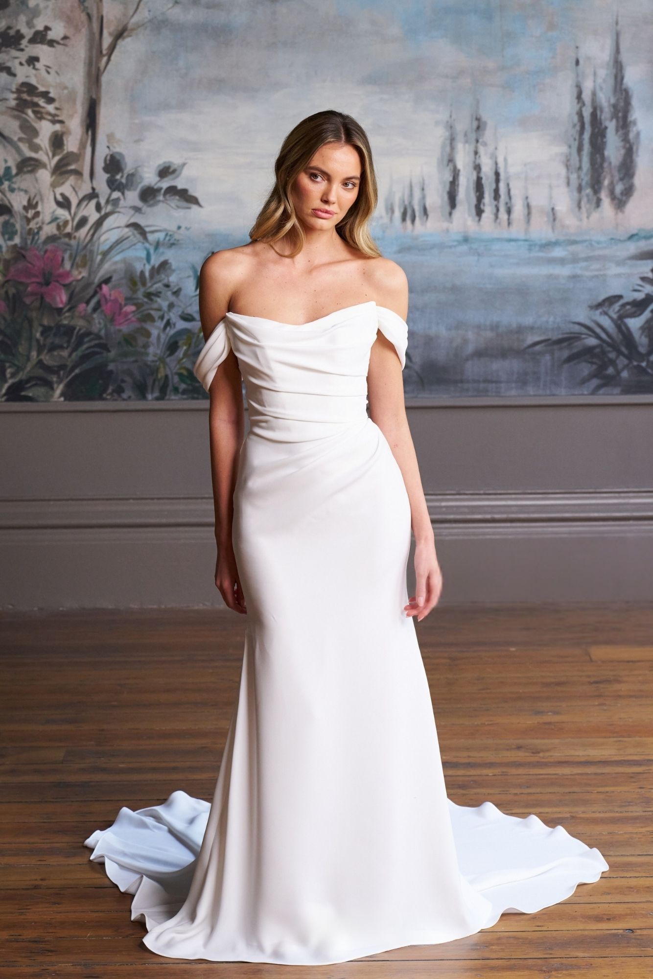A model wearing a white, off-the-shoulder, floor-length bridal gown with a small train against a mural background.