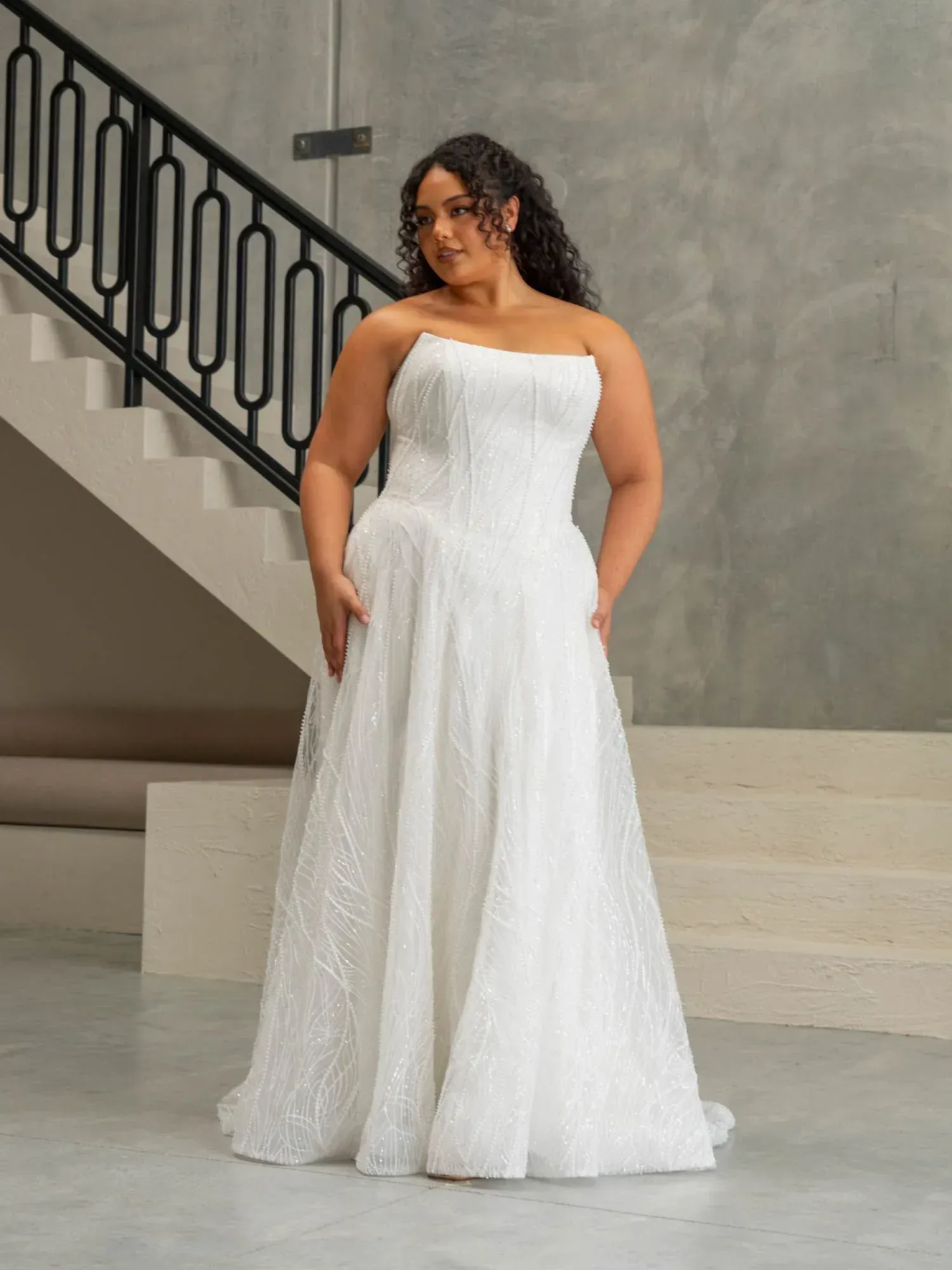 A model stands in a bright, modern indoor space wearing a strapless, floor-length white gown with a textured fabric.