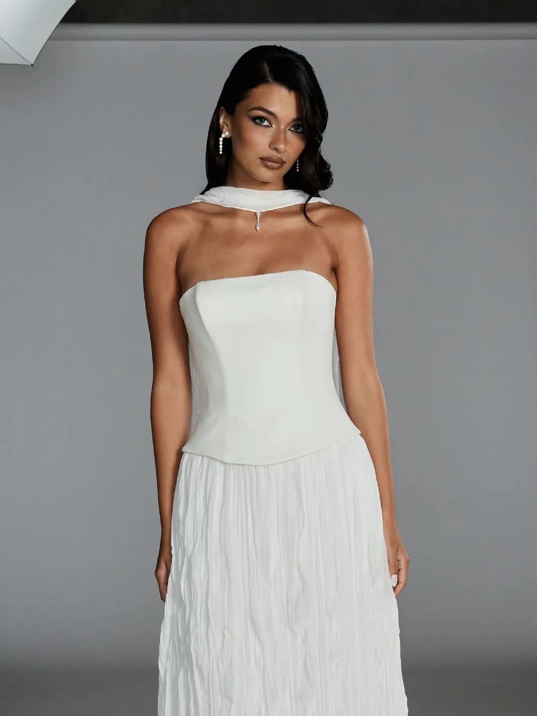 A person wears a strapless, white, corseted gown with a high-neck feather collar against a grey background.