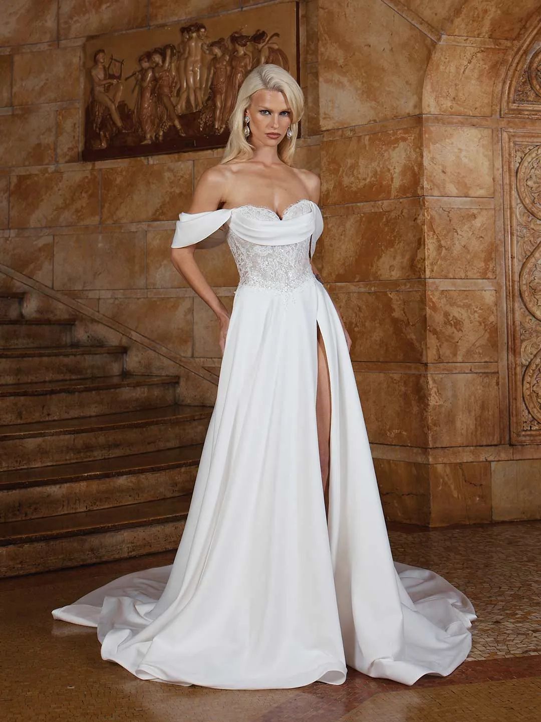 A white off-the-shoulder wedding dress with a lace bodice and a high slit skirt, posed in a stone-walled hallway.