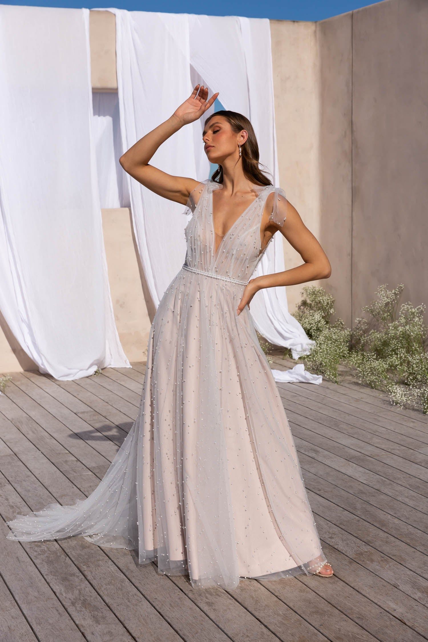 A model wears a floor-length, embellished V-neck gown with a light train, posed outdoors against a draped fabric backdrop.