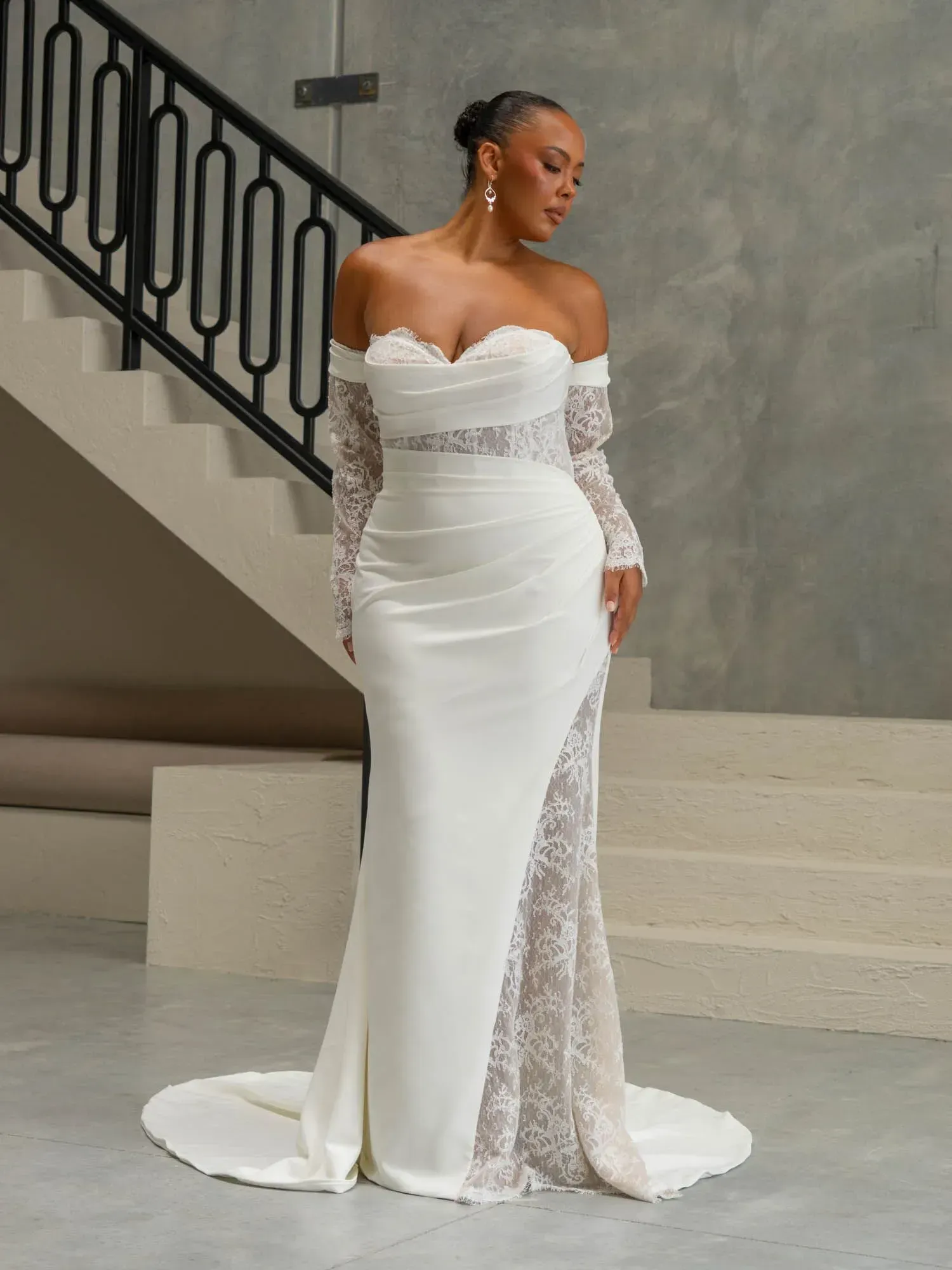 A person modeling an off-the-shoulder, white bridal gown with lace long sleeves and a lace-paneled skirt, posing on stairs.