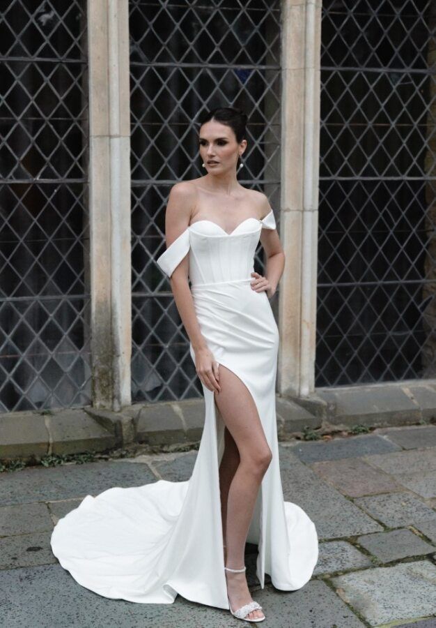 A person wearing a white, off-the-shoulder, floor-length gown with a high slit, standing before a stone-walled window.