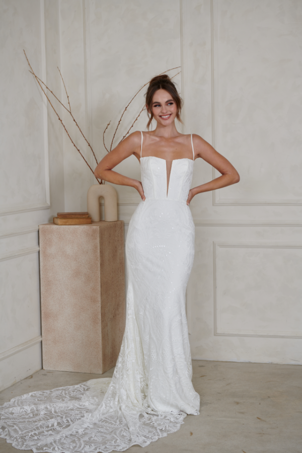 A person wearing a white, floor-length mermaid bridal gown with a deep-v neckline and lace train, posing in a studio.