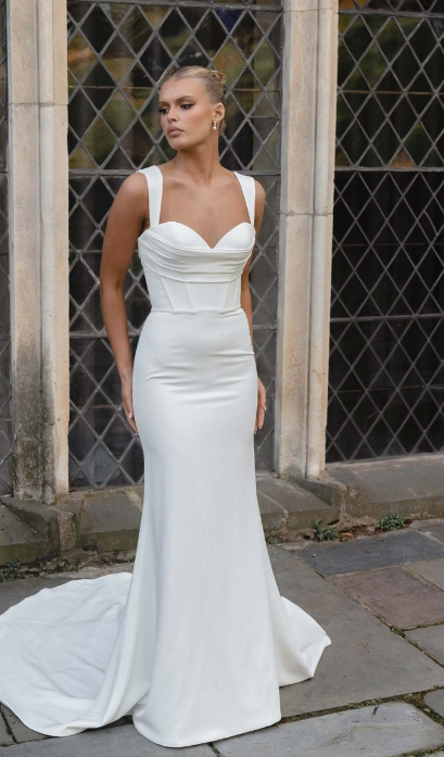 A person wears a white, floor-length, corset-style wedding gown with thick straps, posing outdoors in front of a window.