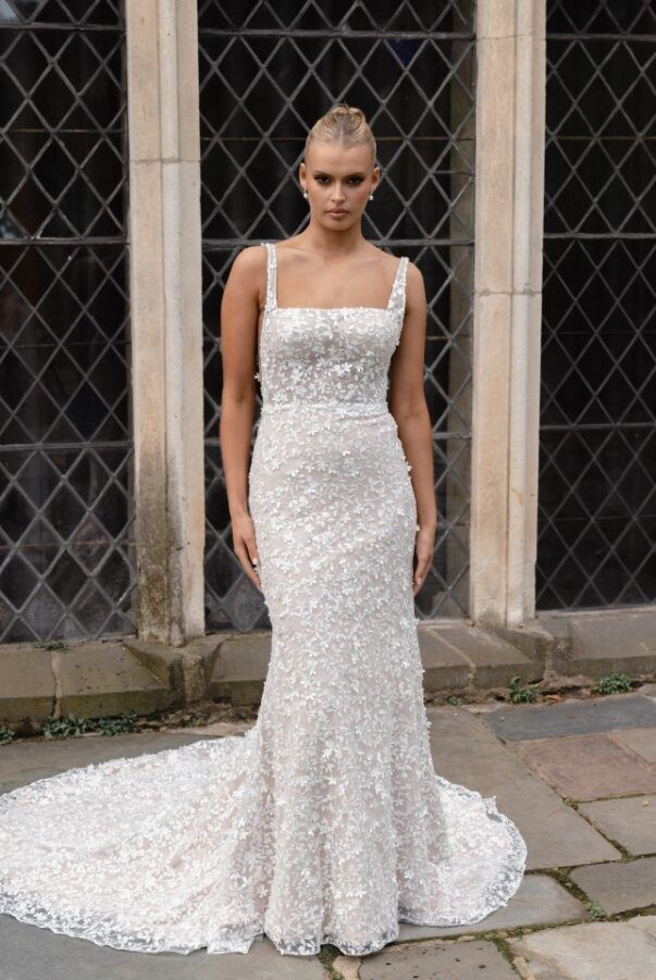 A person in a form-fitting, white, heavily beaded, floor-length wedding dress with a train, standing before stone windows.