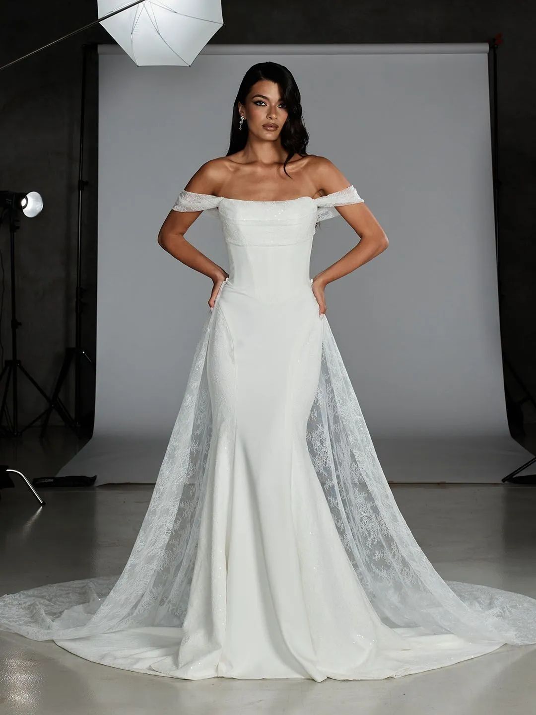 A person in a white, off-the-shoulder mermaid wedding dress with a sheer lace overskirt in a professional photo studio.