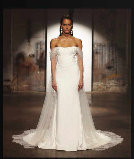 A model wears an off-the-shoulder white wedding dress with sheer tulle sleeves and a floor-length train on a runway.