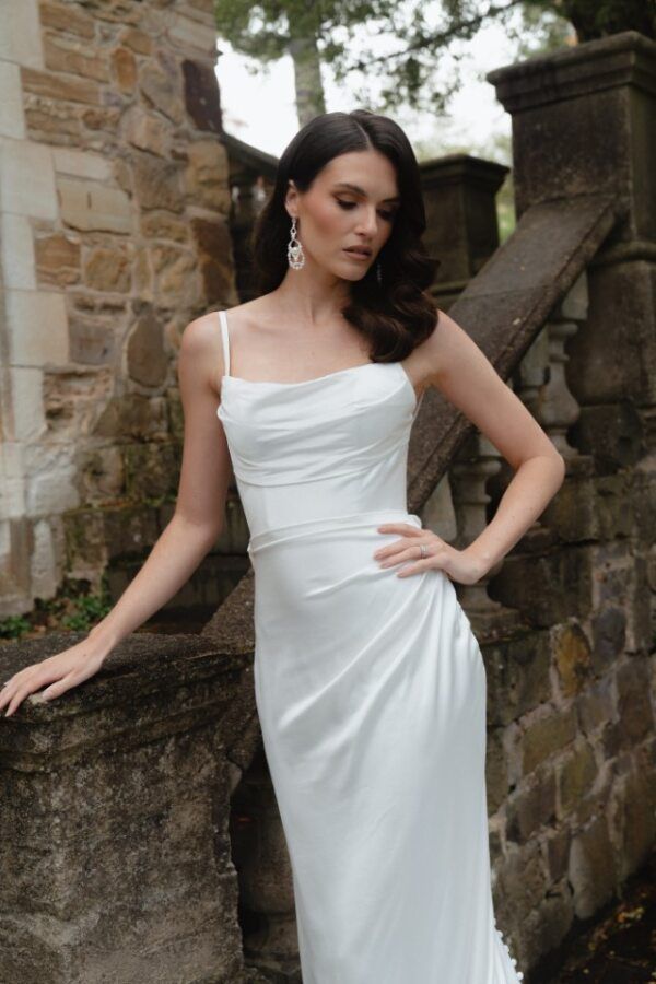 A person in a white, cowl-neck satin slip dress poses against a stone wall in an outdoor, garden-like setting.