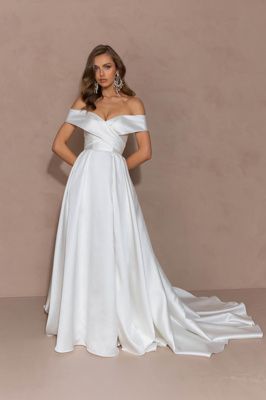 A woman wearing an off-the-shoulder, white satin A-line bridal gown with a floor-length train against a tan background.