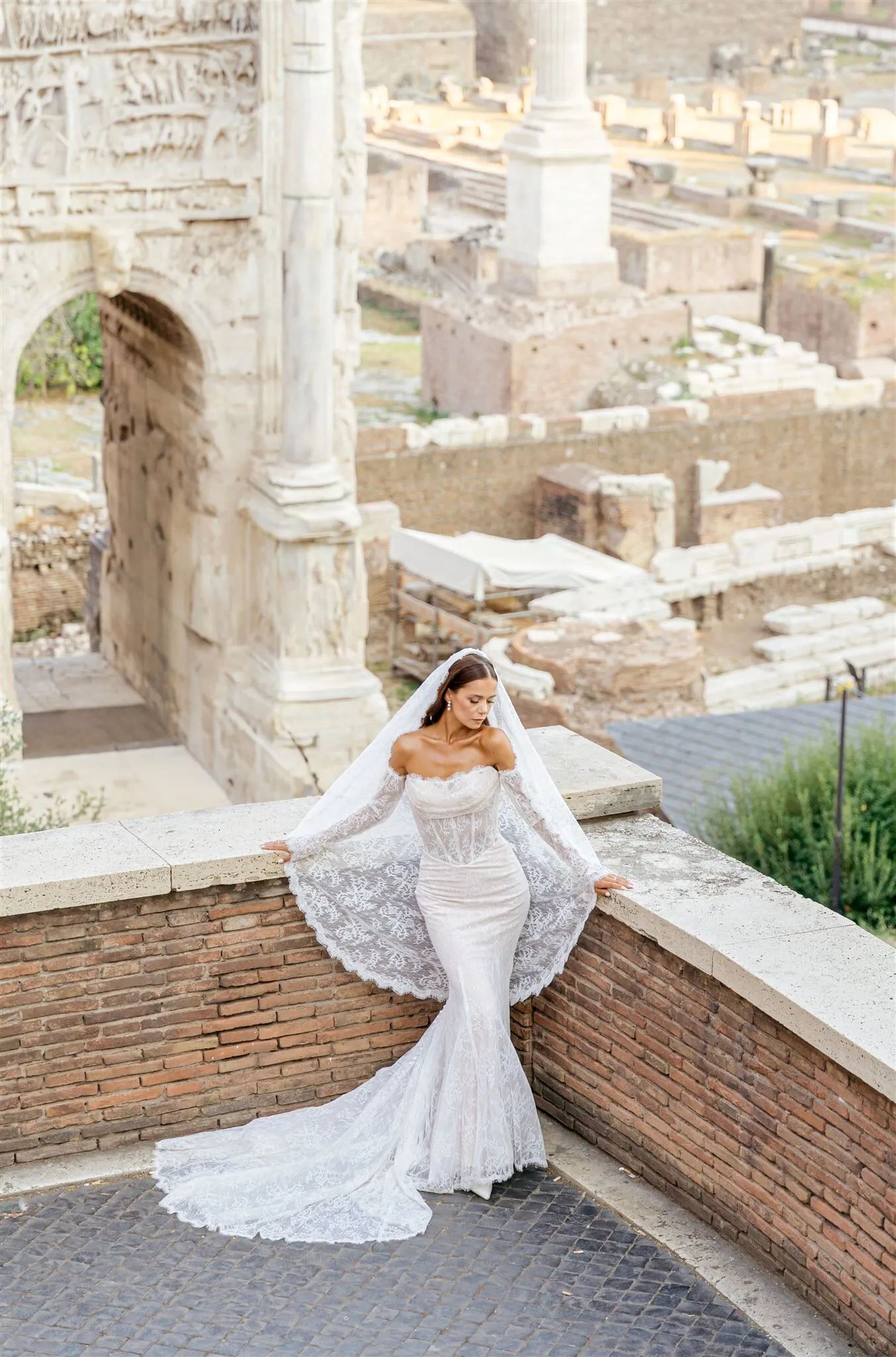 A person in a long, white lace wedding gown and veil poses against a stone archway amidst ancient Roman ruins.