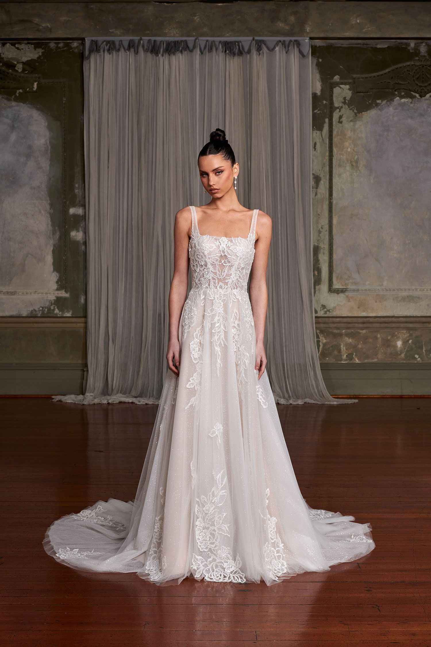 A model wearing an intricately embellished white sleeveless A-line wedding dress with a train in a rustic room.