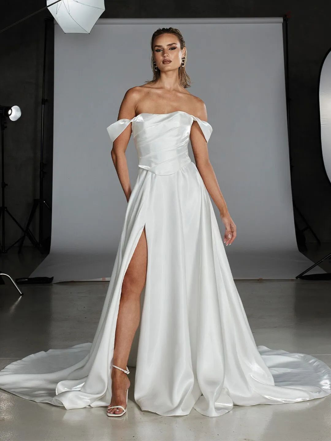 A person wearing a white, satin, off-the-shoulder wedding dress with a high leg slit, posed in a studio setting.
