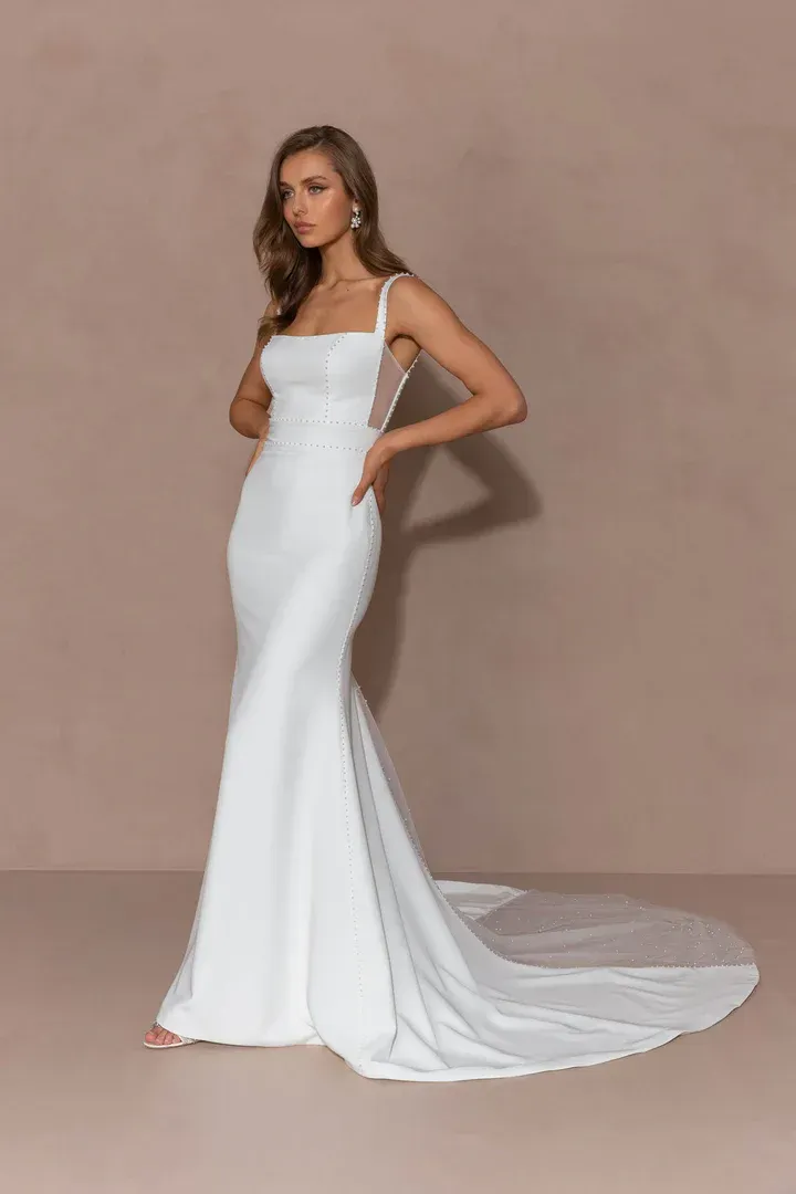 A model in a white, floor-length, square-neck bridal gown with a side cutout and a long train against a beige background.