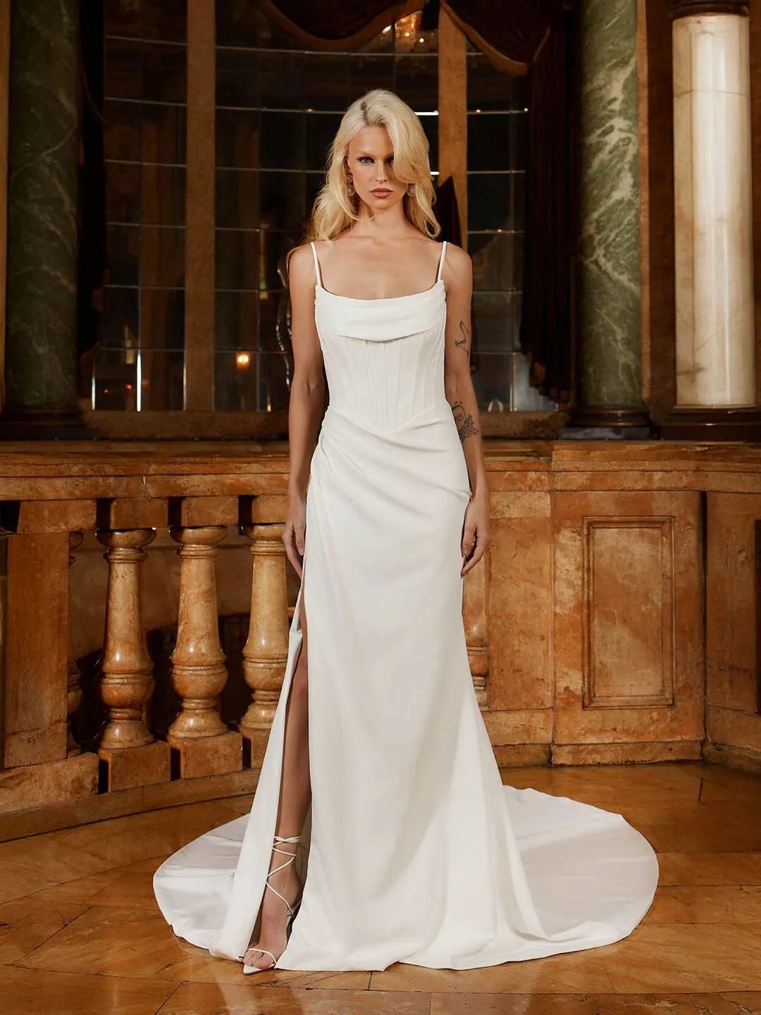 A blonde person wears a white, floor-length, cowl-neck gown with a high thigh slit, standing in an ornate interior.