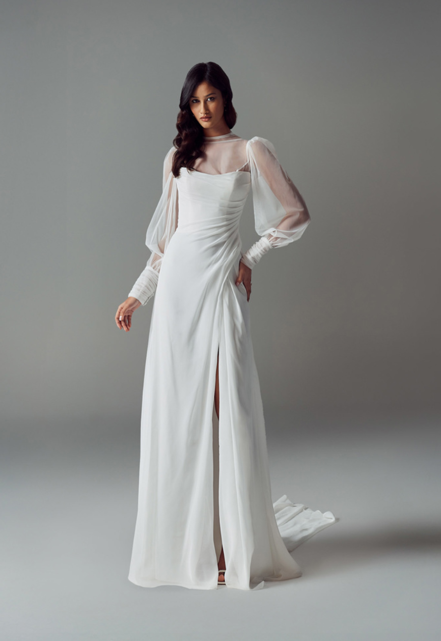 A person models a white, floor-length wedding dress with sheer bishop sleeves, a ruched bodice, and a front leg slit.