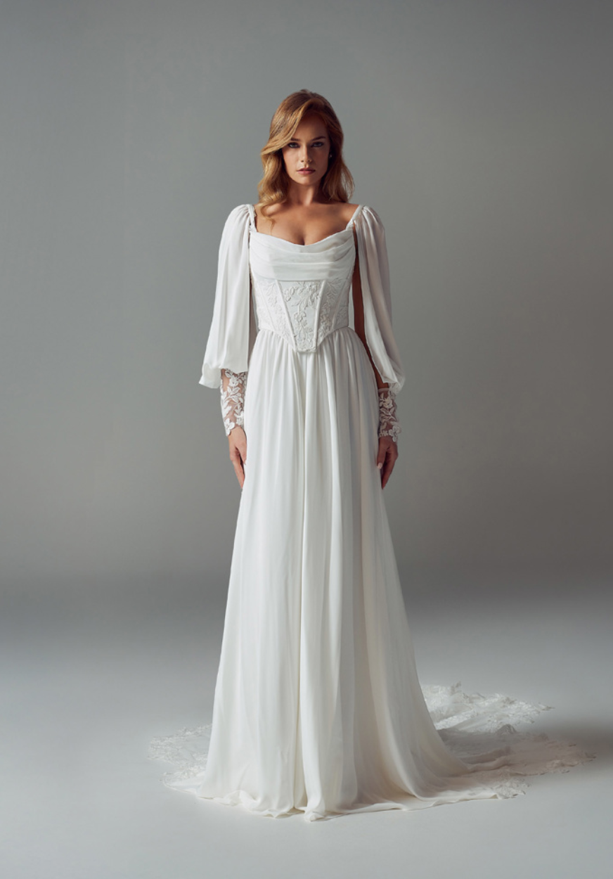 A woman wearing a long-sleeved, white, flowing wedding gown with a corset-style bodice stands against a grey background.