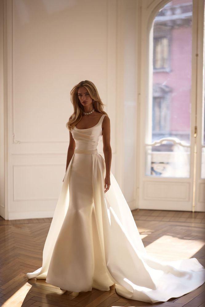 A person wearing a sleek, floor-length white gown with a detachable overskirt, standing in a sunlit room.