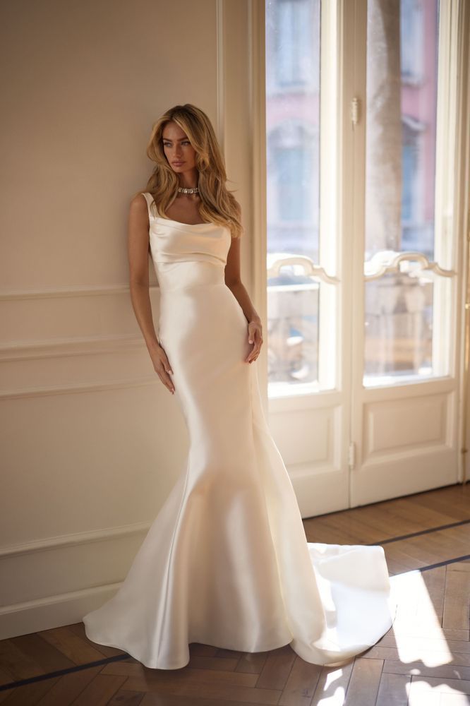 A model wears a form-fitting, sleeveless white satin trumpet gown with a cowl neckline against an interior wall.