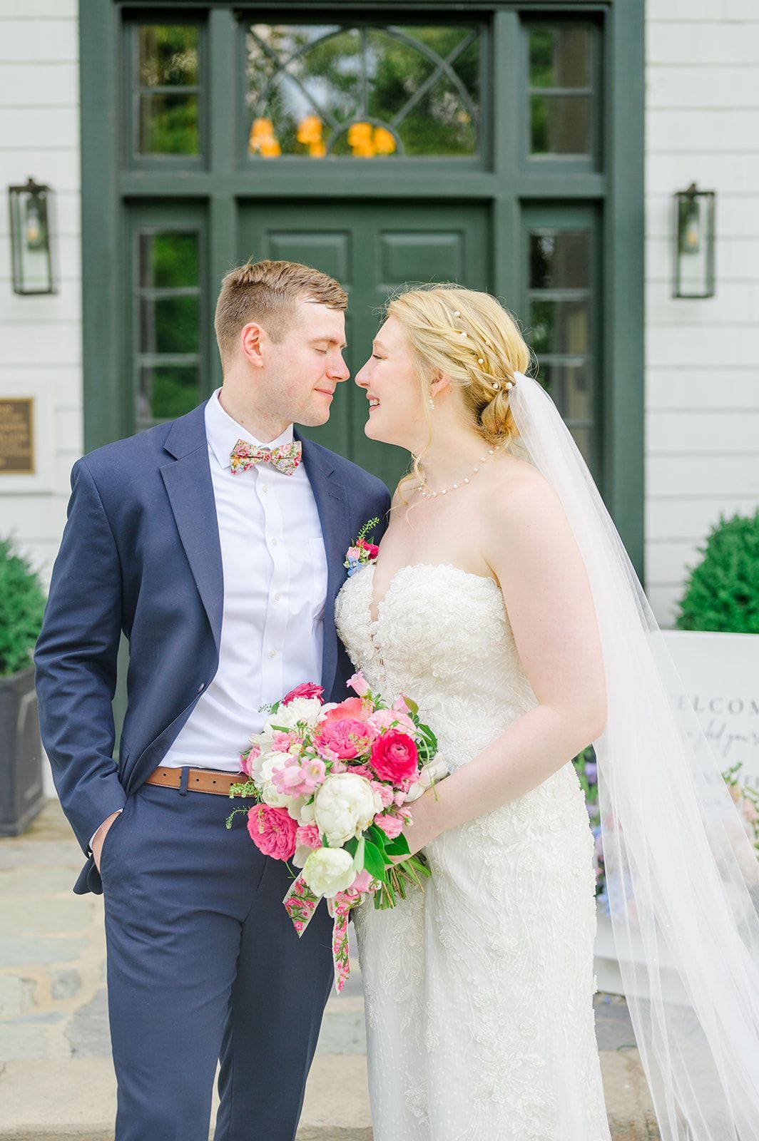 A bride and groom in wedding attire stand close, facing each other with smiles, holding a colorful floral bouquet.