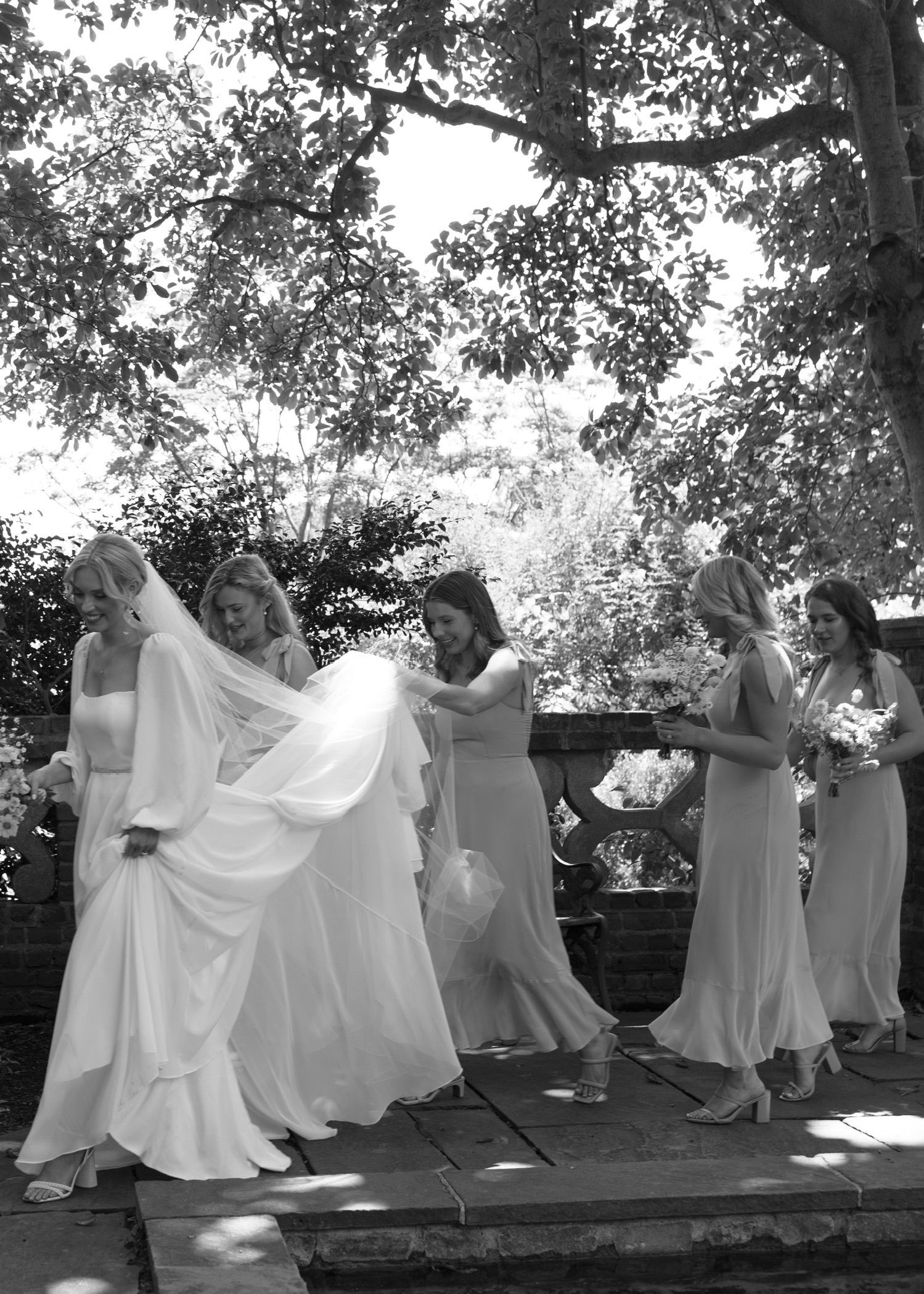 A bride in a long, flowing white gown walks outdoors with four bridesmaids in light-colored dresses following behind.