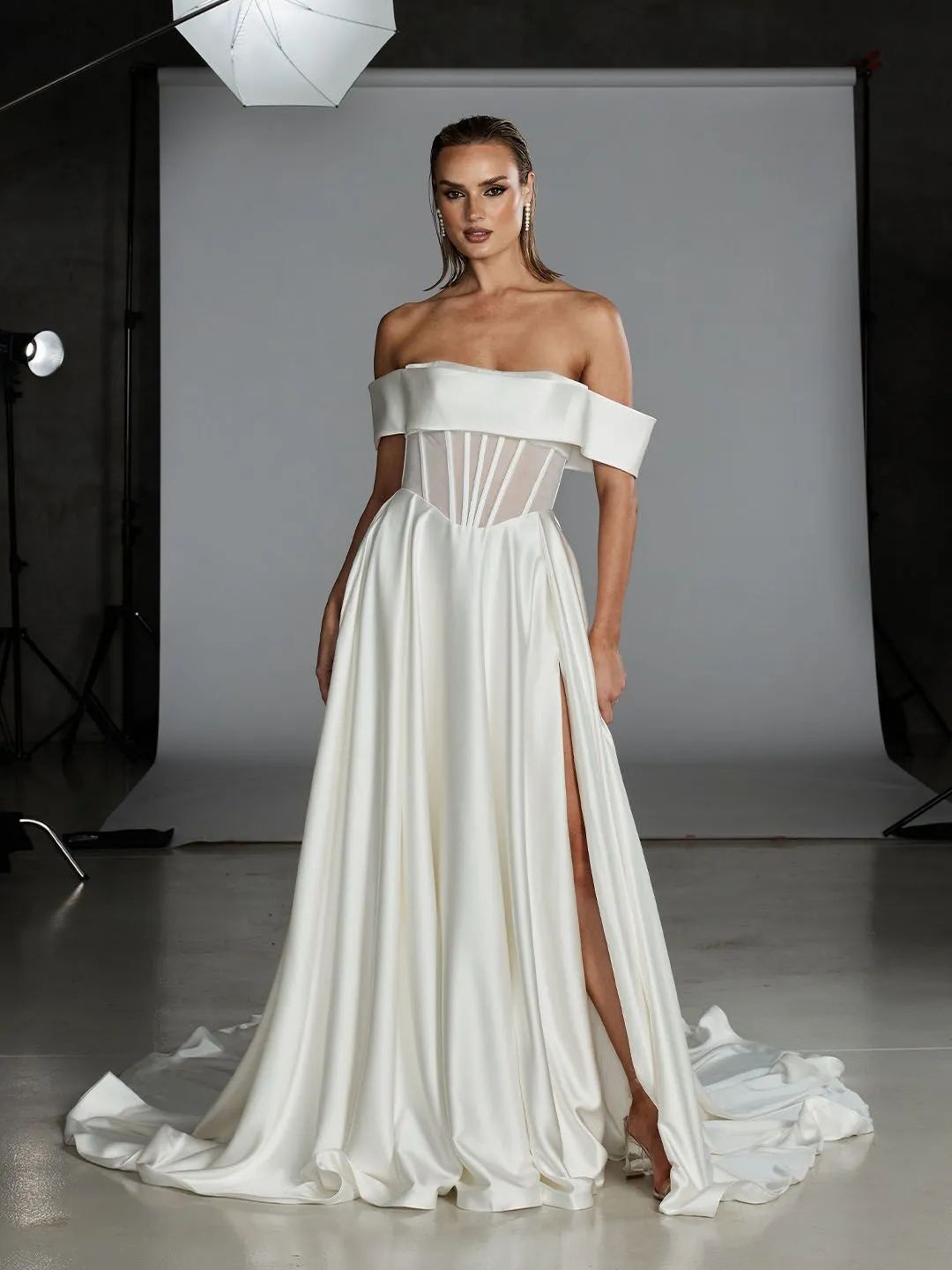 A model poses in a white, off-the-shoulder wedding gown with a sheer corset bodice and a high slit in a photography studio.