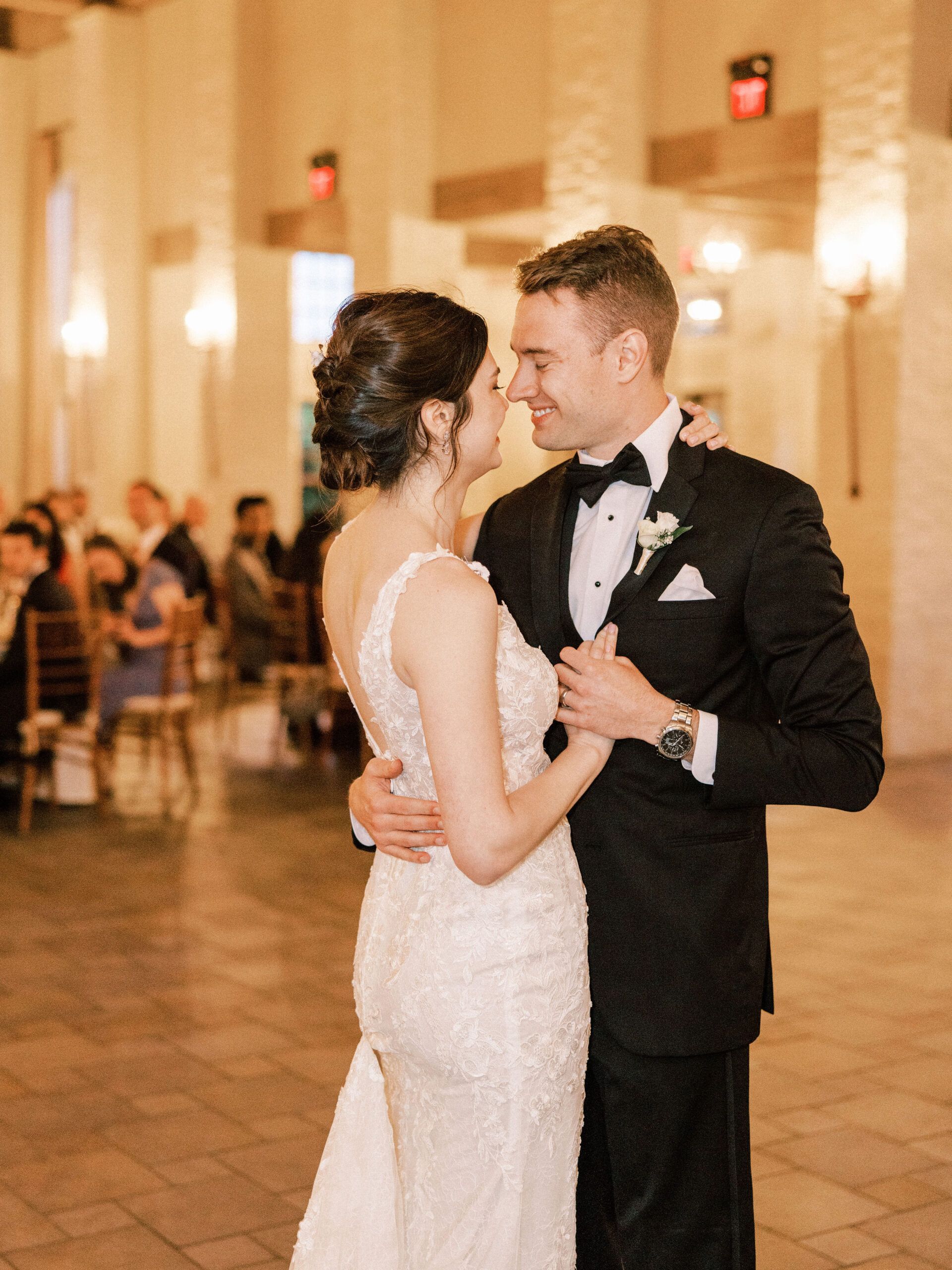 A couple in formal attire dances together in a brightly lit ballroom, gazing at each other with smiles.