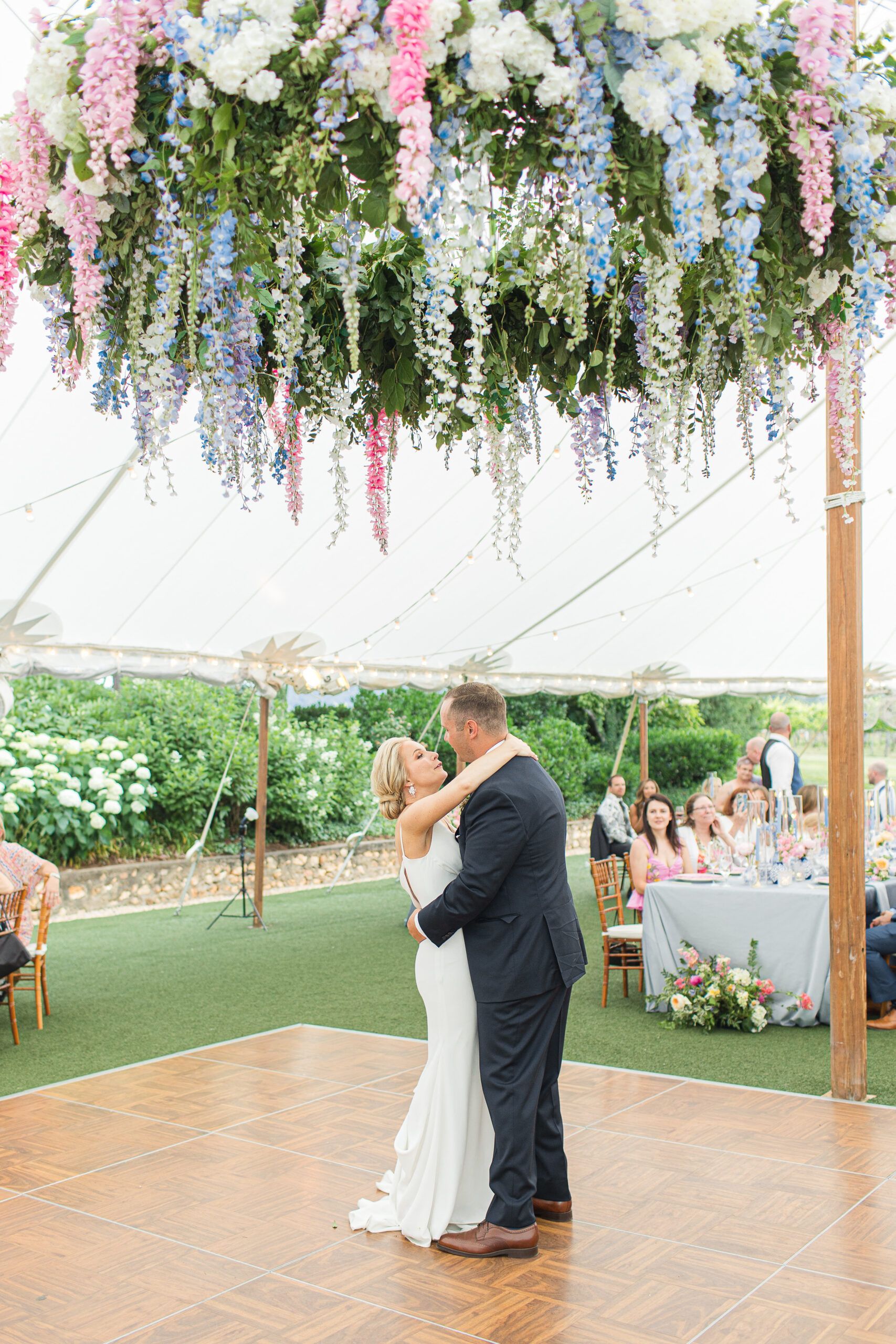 A couple dances on a wooden floor under a large, vibrant floral chandelier in an outdoor tented wedding venue.