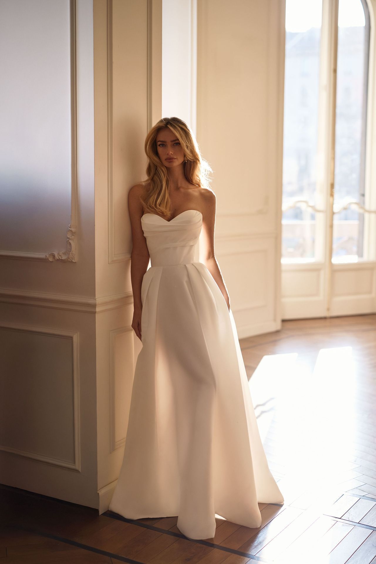 A person with wavy hair wearing a strapless white A-line gown, leaning against a white pillar in a sunlit room.