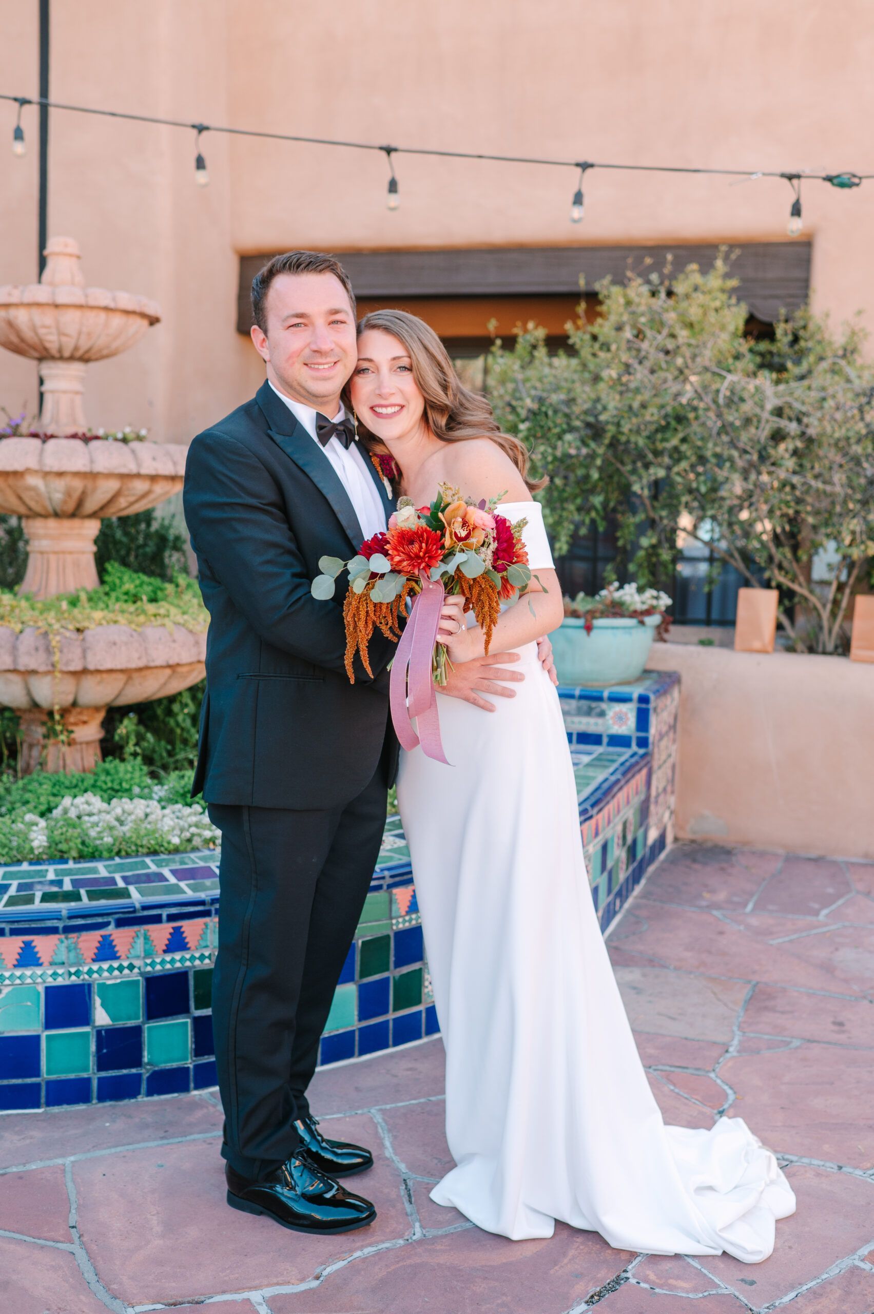 A couple in wedding attire poses together holding a vibrant floral bouquet by an outdoor tiered fountain and tile wall.