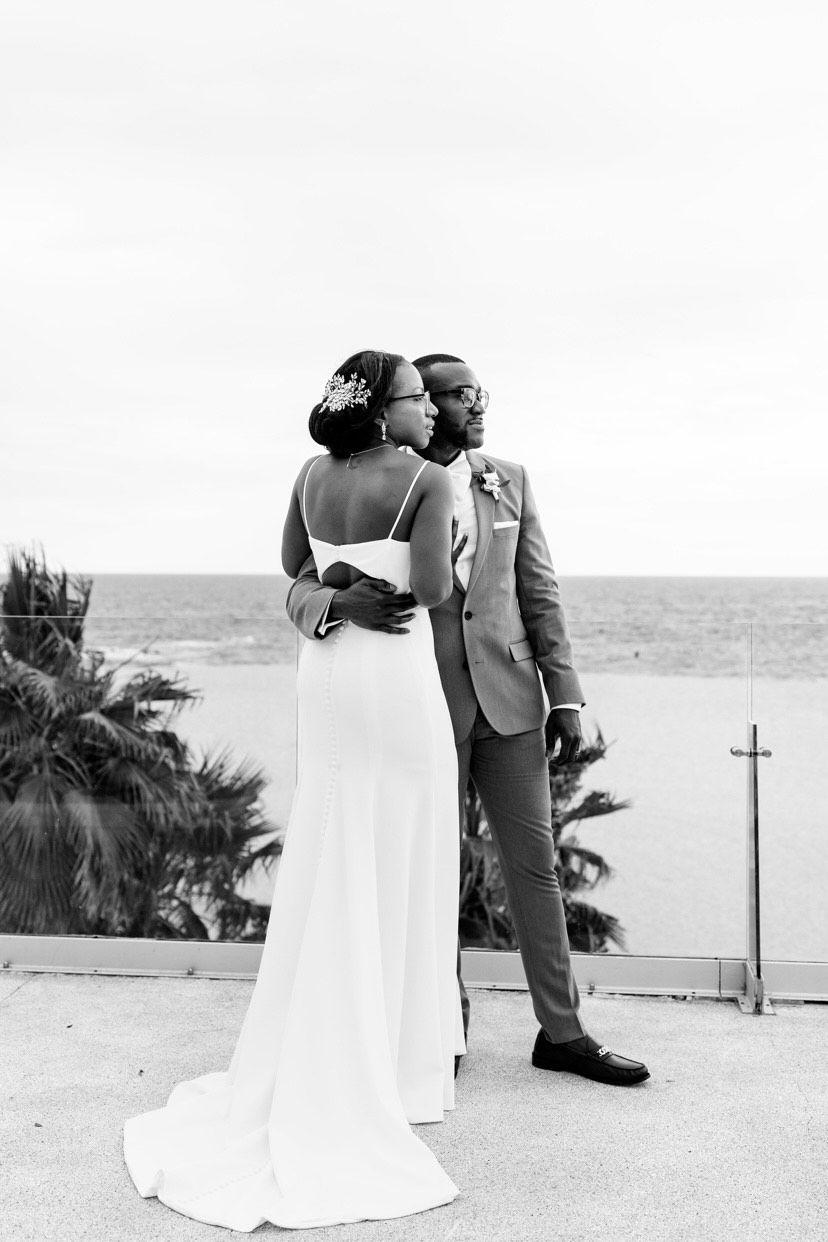 A couple embraces while standing on a balcony overlooking the ocean in a black-and-white photo.