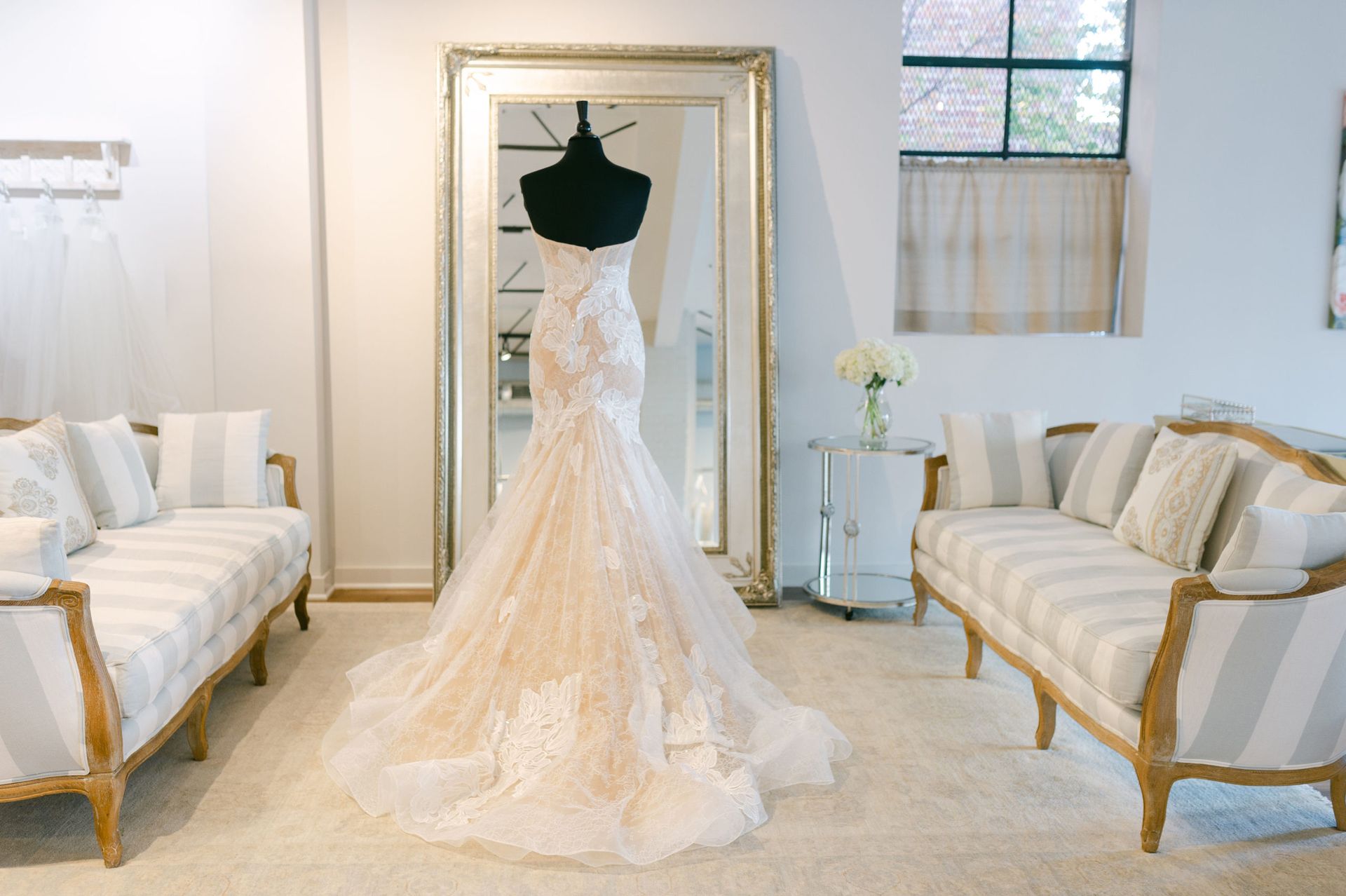 A white lace, mermaid-style wedding dress on a mannequin, centered between two striped sofas in a bright boutique.