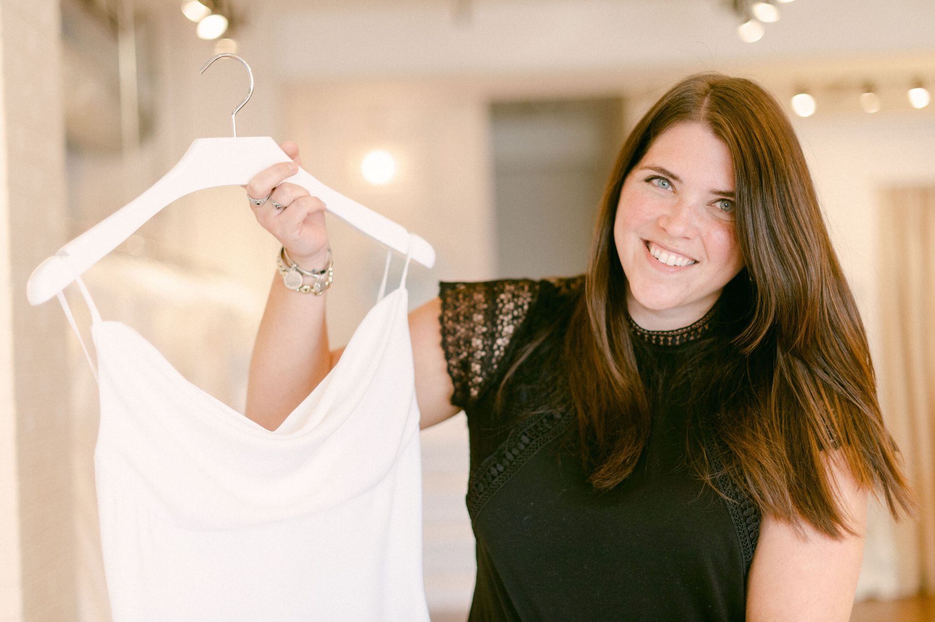A smiling person in a black lace-sleeved top holds up a white bridal gown on a hanger in a bright, modern shop.