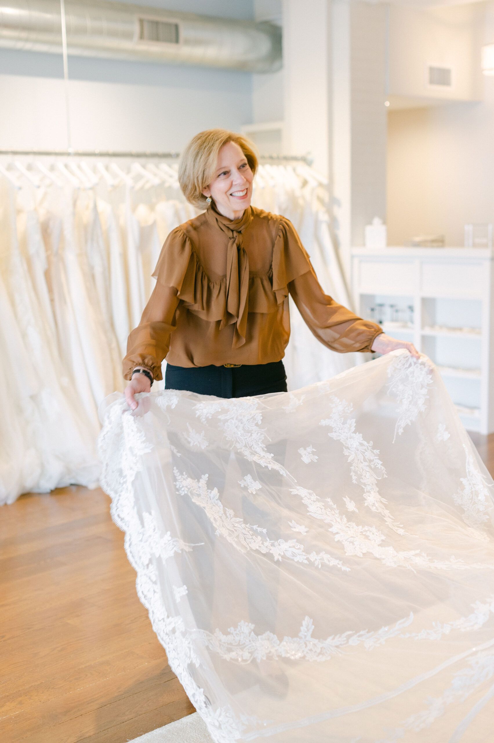 A person in a brown ruffled blouse holds up a long, white, lace-trimmed bridal veil in a dress boutique.