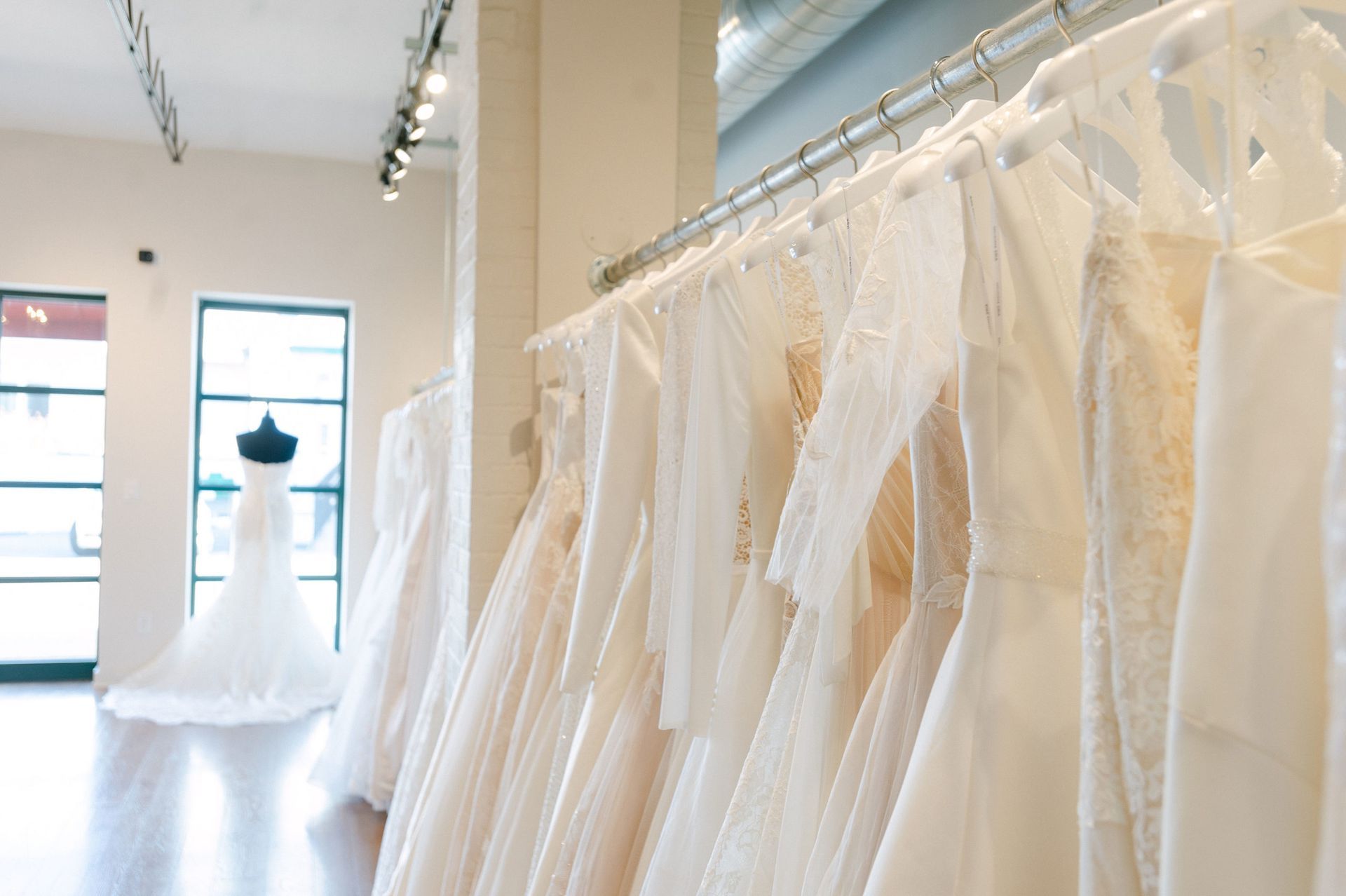 A row of white wedding dresses hanging on a rack in a bright boutique, with a mannequin in a gown in the background.